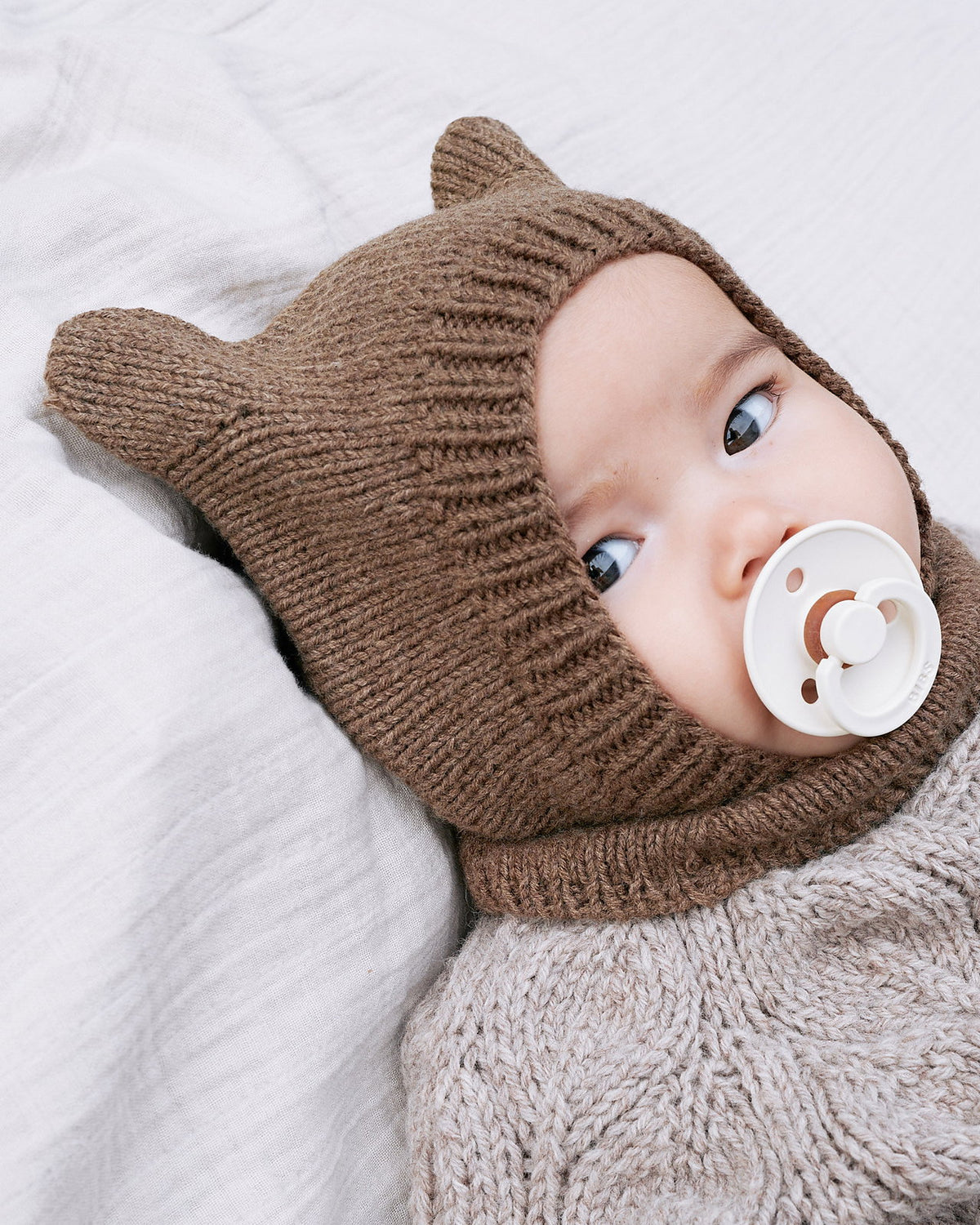 Brown knitted bear-ear hood with a ribbed face band worn by a baby